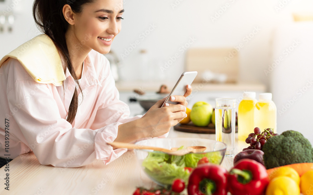 Smiling young woman using cellphone while cooking dinner in kitchen at home, cheerful casual female leaning on table, reading text messages on her cell phone and smiling, free copy space Smiling young woman using cellphone while cooking dinner in kitchen at home, cheerful casual female leaning on table, reading text messages on her cell phone and smiling, free copy space