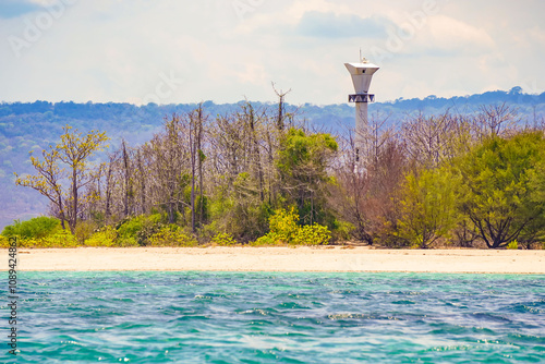 A serene coastal landscape with a lighthouse standing tall amidst a cluster of trees on a small island. The clear blue water gently laps against the sandy shore, creating a picture-perfect scene. photo