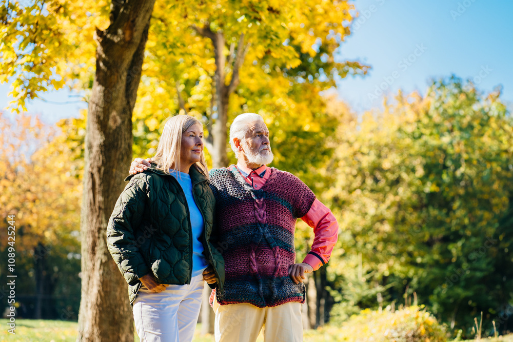 Fototapeta premium Older couple, man and woman, stroll through park surrounded by vibrant autumn colors with trees with golden leaves form beautiful backdrop for peaceful, leisurely walk.