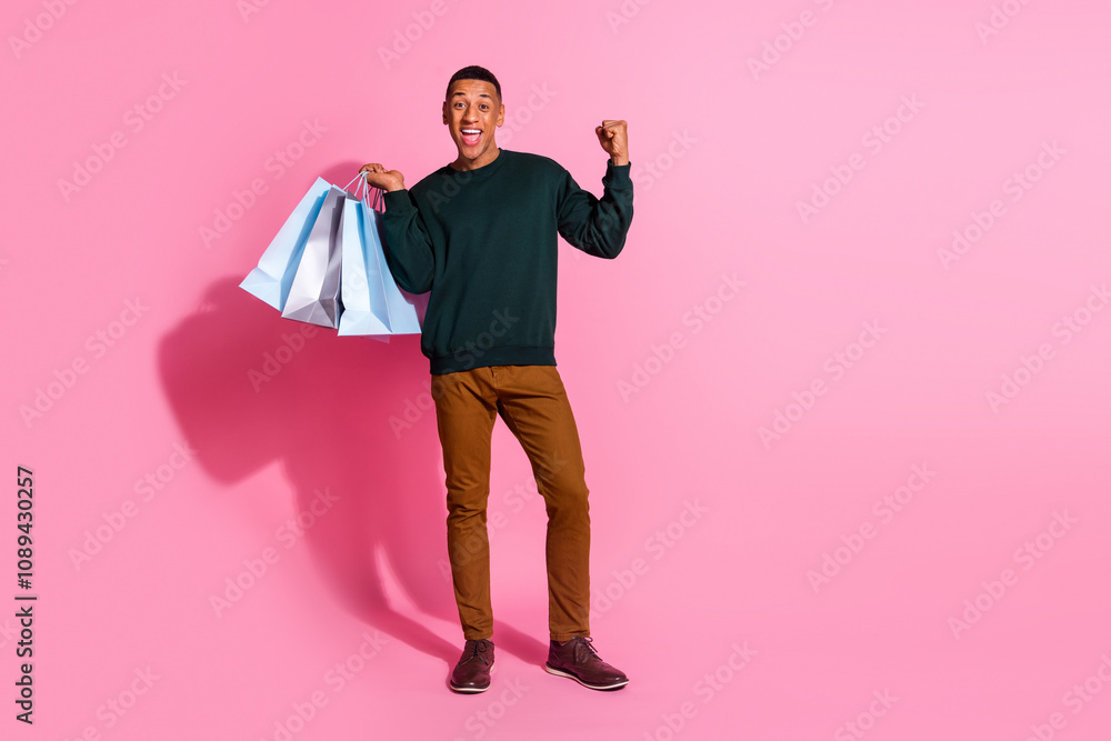 Full length photo of lucky positive guy dressed pullover rising fist holding bargains emtpy space isolated pink color background Full length photo of lucky positive guy dressed pullover rising fist holding bargains emtpy space isolated pink color background