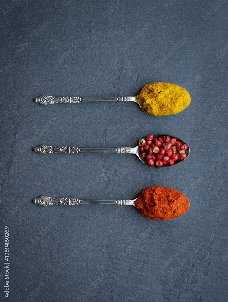 Three Silver Tea Spoons with turmeric, red peppercorns and chilli powder on a grey slate background. Three Silver Tea Spoons with turmeric, red peppercorns and chilli powder on a grey slate background.