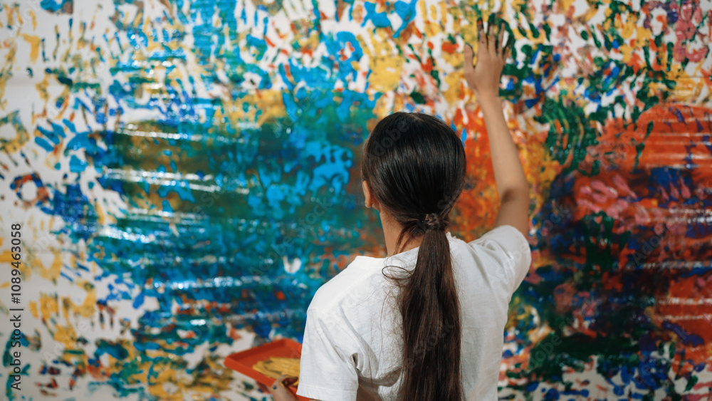 Obraz premium Young happy girl holding the color tray and paint colorful stained wall to express idea and enhance imagination with hand print. Caucasian girl stamp hand to decorate room in art lesson. Edification.