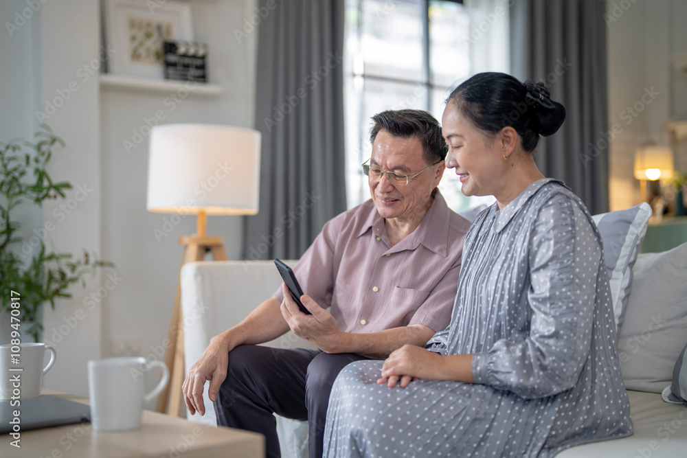 An elderly couple sits together in their cozy living room, smiling and enjoying a moment as they look at something on a smartphone. Their warmth and affection capture a peaceful home atmosphere. An elderly couple sits together in their cozy living room, smiling and enjoying a moment as they look at something on a smartphone. Their warmth and affection capture a peaceful home atmosphere.