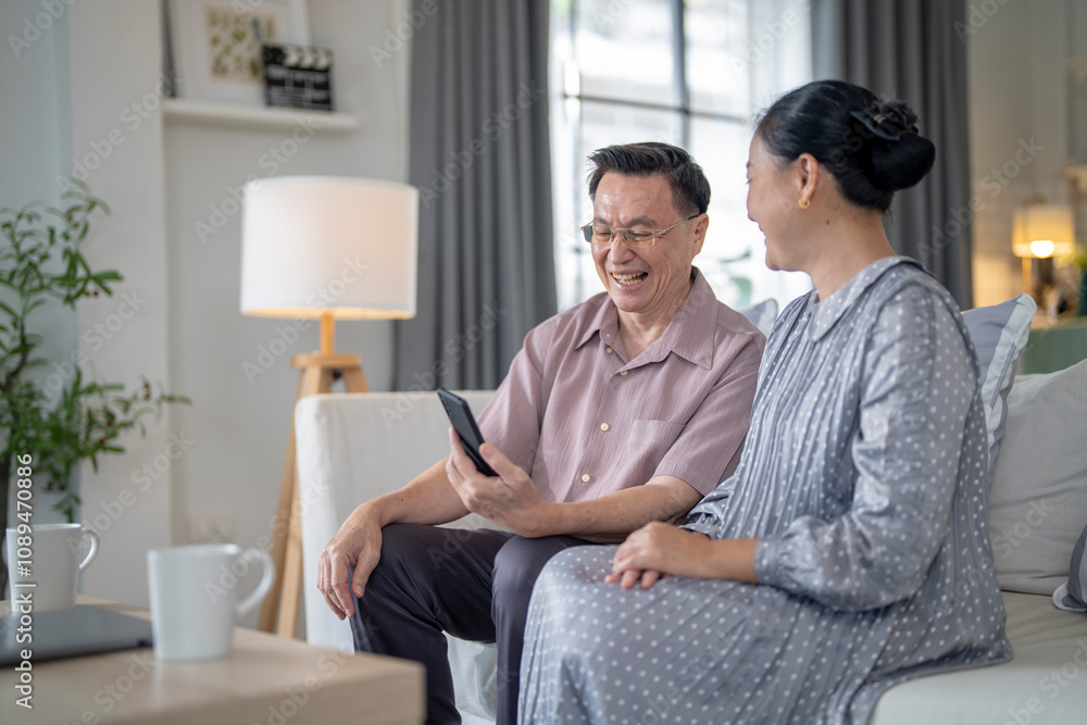 An elderly couple sits together in their cozy living room, smiling and enjoying a moment as they look at something on a smartphone. Their warmth and affection capture a peaceful home atmosphere. An elderly couple sits together in their cozy living room, smiling and enjoying a moment as they look at something on a smartphone. Their warmth and affection capture a peaceful home atmosphere.