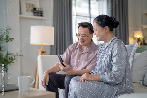 An elderly couple sits together in their cozy living room, smiling and enjoying a moment as they look at something on a smartphone. Their warmth and affection capture a peaceful home atmosphere.