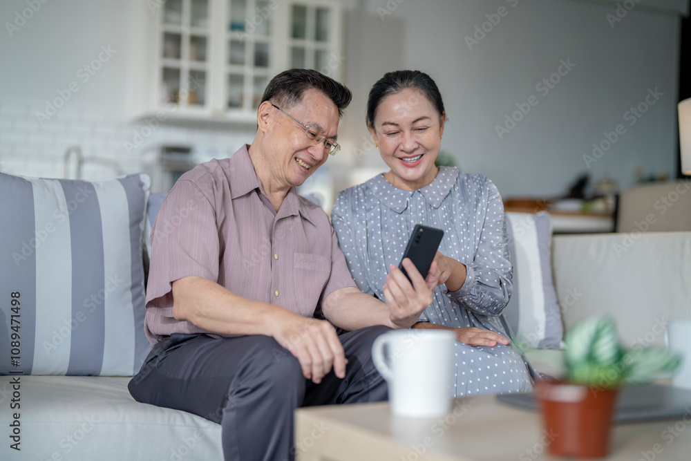 An elderly couple sits together in their cozy living room, smiling and enjoying a moment as they look at something on a smartphone. Their warmth and affection capture a peaceful home atmosphere. An elderly couple sits together in their cozy living room, smiling and enjoying a moment as they look at something on a smartphone. Their warmth and affection capture a peaceful home atmosphere.