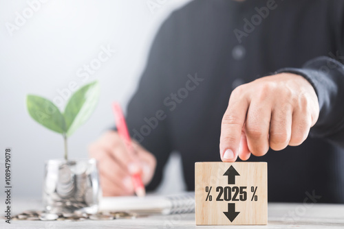 Businessman points at wooden blocks while a coin jar with a sprouting plant sits nearby. He notes numbers in a notebook, symbolizing analysis of 2025 interest rate trends-rise or fall