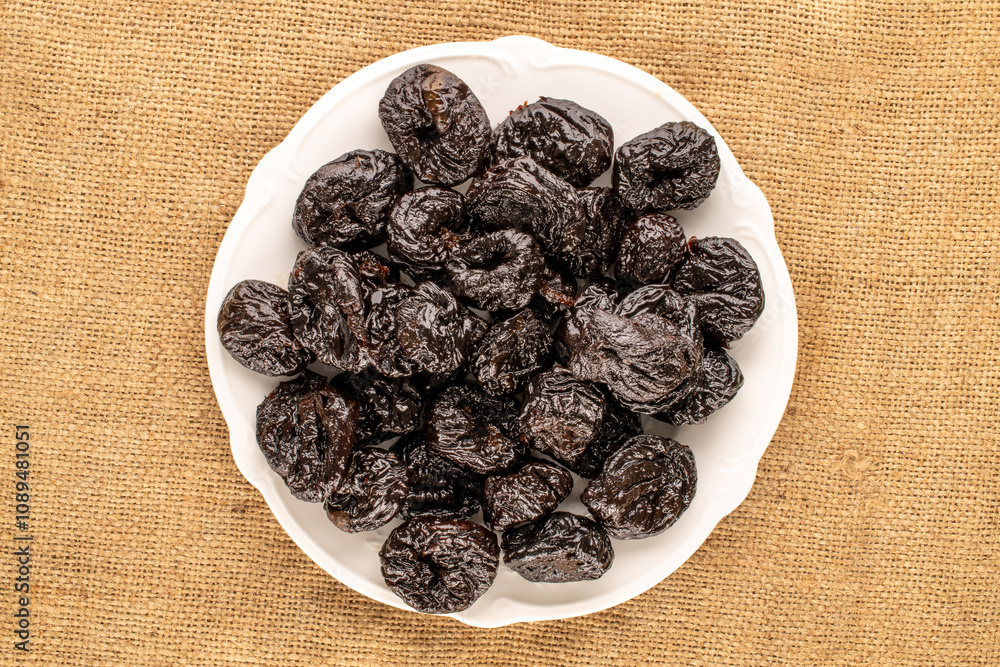 Sweet prunes in a ceramic plate on a jute cloth, macro, top view. Sweet prunes in a ceramic plate on a jute cloth, macro, top view.