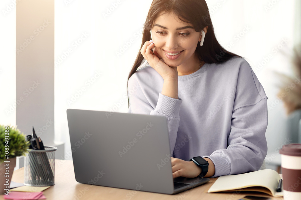 People And Modern Technology Concept. Portrait of smiling woman in wireless headphones sitting at desk, using laptop and watching tutorial, lecture or webinar, studying online at home People And Modern Technology Concept. Portrait of smiling woman in wireless headphones sitting at desk, using laptop and watching tutorial, lecture or webinar, studying online at home