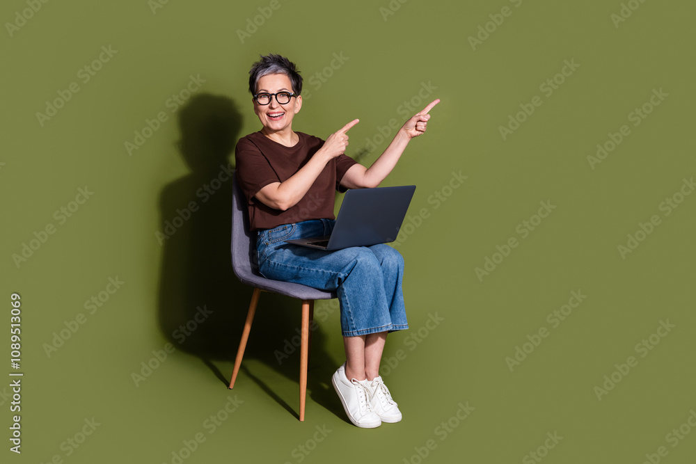Full length photo of attractive retired woman sit chair netbook point empty space dressed brown clothes isolated on khaki color background Full length photo of attractive retired woman sit chair netbook point empty space dressed brown clothes isolated on khaki color background