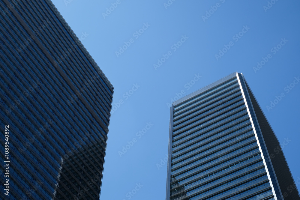 A low-angle view of two modern skyscrapers with glass facades under a clear blue sky, emphasizing contemporary architecture and urban development in a cityscape. A low-angle view of two modern skyscrapers with glass facades under a clear blue sky, emphasizing contemporary architecture and urban development in a cityscape.