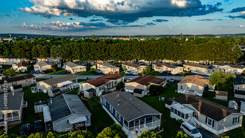 Aerial view captures a Mobile, Prefab, Manufactured Home neighborhood with houses surrounded by tall trees. The sun sets in the background, casting a warm glow over the rooftops lush landscape.