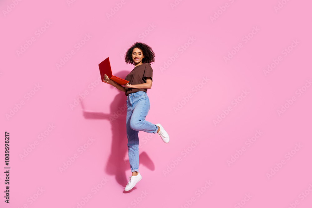 Full length photo of lovely young lady jump hold netbook dressed stylish brown garment isolated on pink color background Full length photo of lovely young lady jump hold netbook dressed stylish brown garment isolated on pink color background