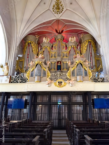 A detailed low-angle view of the historic pipe organ in Konigsberg Cathedral. The baroque design features intricate gold ornamentation and angelic sculptures, framed by soaring gothic arches and photo