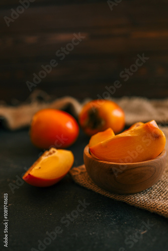 Rustic composition with fresh persimmons in a wooden bowl and on a dark textured background, accented by burlap. Ideal for seasonal, culinary, and rustic lifestyle themes photo