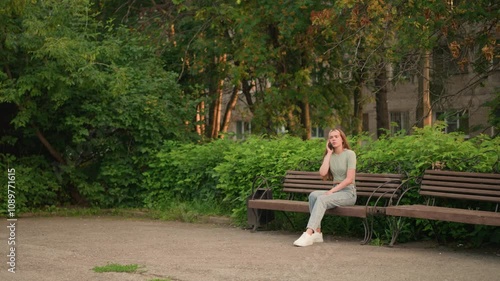 Lady on phone call sits on wooden bench, looking down thoughtfully as she hangs up, surrounded by lush greenery, trees, and a building in background with sunlight filtering through leaves photo