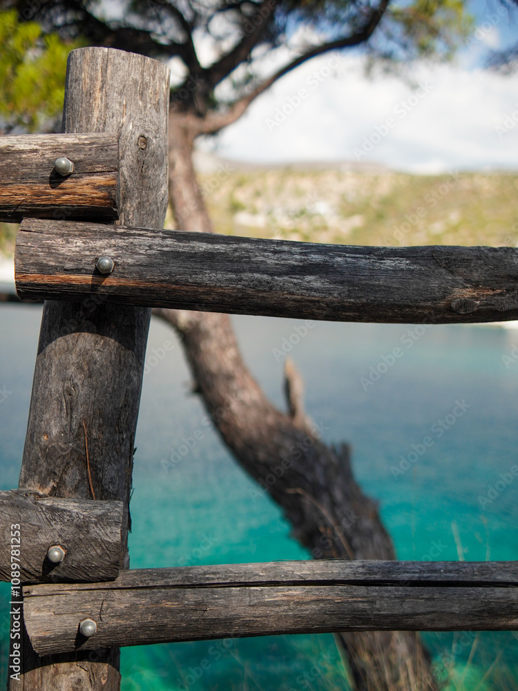 Fototapeta premium Weathered wooden fence posts and rails are fastened together with metal bolts. A blurred background reveals a scenic coastal view featuring clear turquoise water and a dry, scrubby tree.