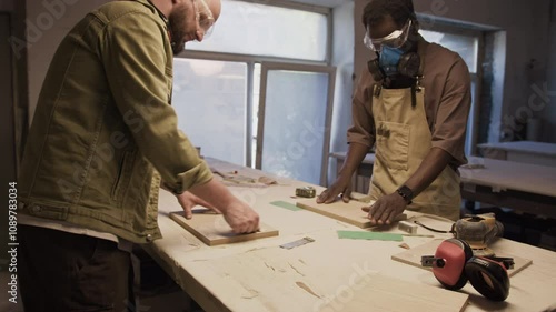 Zoom out shot of African American craftsman in protective respirator mask and goggles discussing material with Caucasian coworker while he sanding wooden board at workbench in carpentry workshop photo