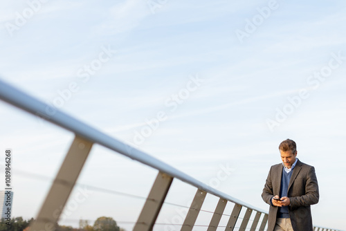 A businessman checks his smartphone while standing on a modern bridge against a clear blue sky during early evening in the city