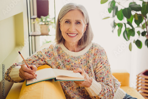 Charming elderly woman enjoying a relaxing weekend at home with a smile, writing in a journal while basking in daylight