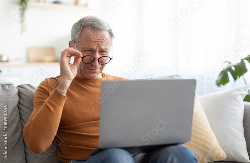 Poor Eyesight. Senior Man Squinting Eyes Using Laptop Wearing Eyeglasses Having Problems With Vision Sitting On Couch. Ophtalmic Issue, Bad Sight In Older Age, Macular Degeneration Concept photo