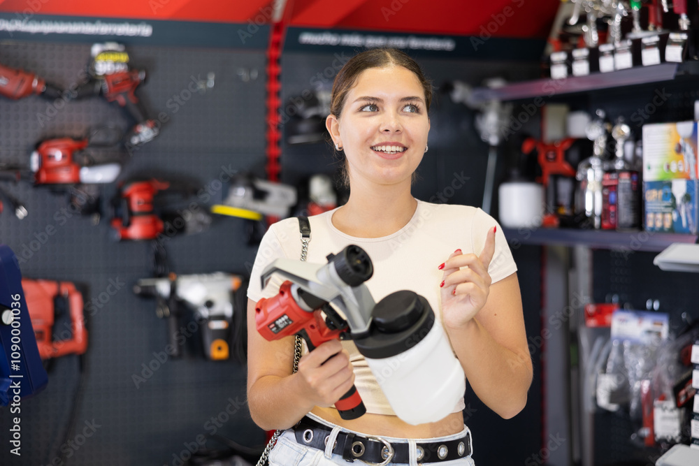 Fototapeta premium Woman examines handheld wireless portable spray gun, airbrush in tools department of construction store. Girl consider house or apartment renovation redecoration. Interior design, styling concept.