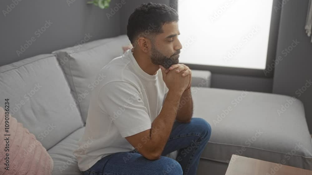 Young man sitting in living room, looking serious and contemplative, dressed casually in a white shirt and jeans, with a modern and cozy apartment background highlighting quiet indoor ambiance Young man sitting in living room, looking serious and contemplative, dressed casually in a white shirt and jeans, with a modern and cozy apartment background highlighting quiet indoor ambiance