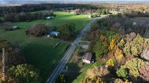 Driving cars on rural road in suburb of American town. Colorful trees and green farm fields in autumn season. Aerial flyover shot. Single family houses and barns in USA. Top down shot.