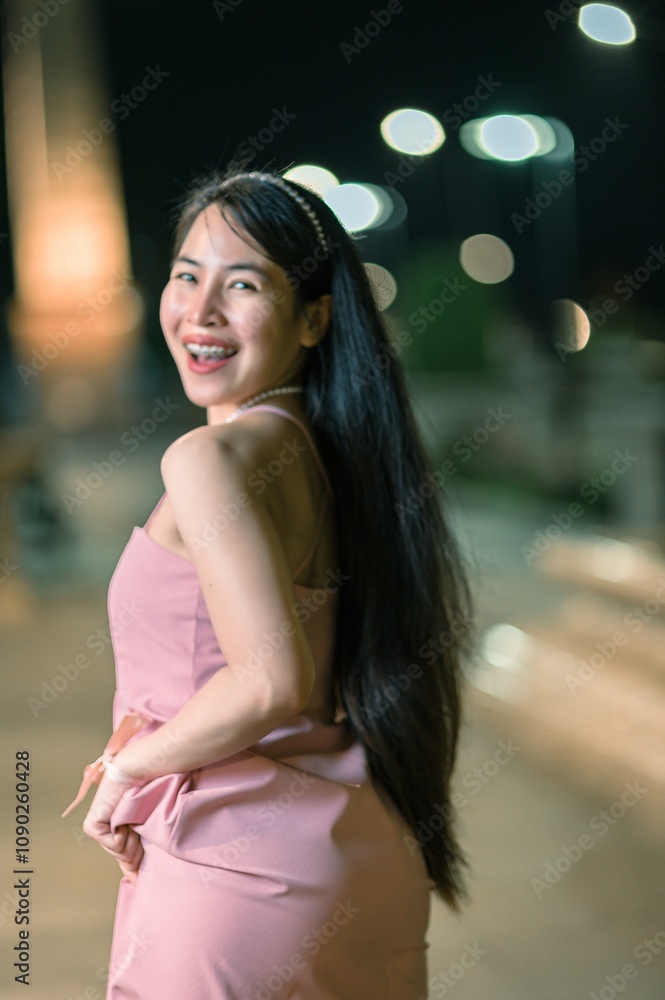 Fototapeta premium Joyful woman in a pink dress poses confidently at a vibrant night setting, showcasing her long hair and radiant smile against a backdrop of glowing lights.