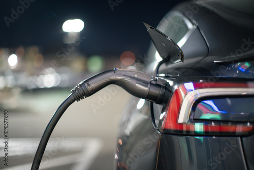 Electric car charging in urban parking lot at night, focus on EV charger station cable plugged into charging port. Closeup.