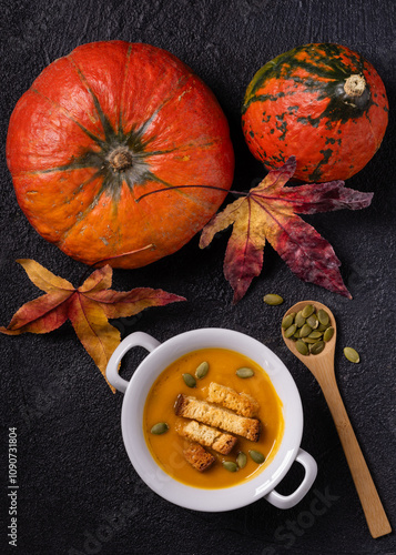 on a textured black background, top view, a white bowl of pumpkin soup garnished with pumpkin seeds and croutons. On the side, pumpkins and autumn leaves. photo