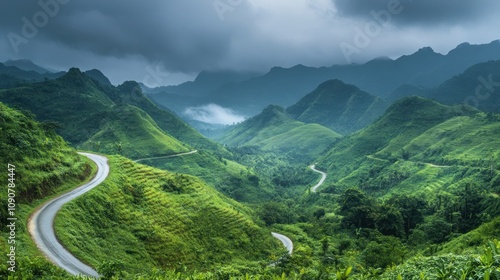 A winding road snakes through a lush green mountain valley with misty mountains in the background under an overcast sky.