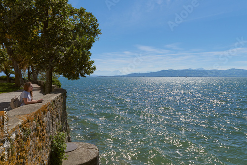 Enchanting Young Woman Admires Bodensee Panorama from Medieval Castle Walls in Lindau, Germany. Idyllic Summer Scene Captures European Vacation Bliss with Alpine Mountains, Yacht