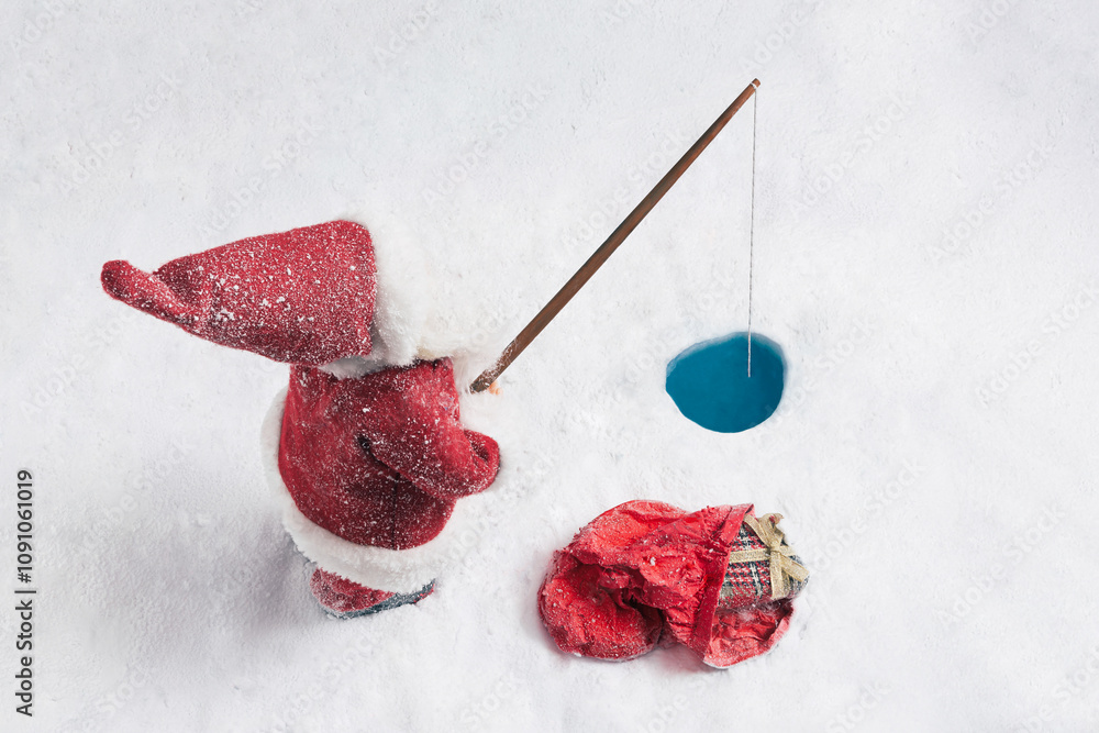 A snow-covered Santa Claus figure ice fishes on a frozen lake. A small hole has been cut in the ice, and a fishing rod is cast. His sack of gifts lies nearby in the snow. A snow-covered Santa Claus figure ice fishes on a frozen lake. A small hole has been cut in the ice, and a fishing rod is cast. His sack of gifts lies nearby in the snow.