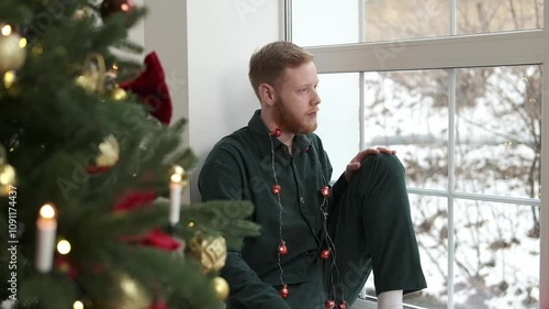 An attractive red-haired, bearded guy is sitting on the windowsill by the Christmas tree.New Year and Christmas concept.