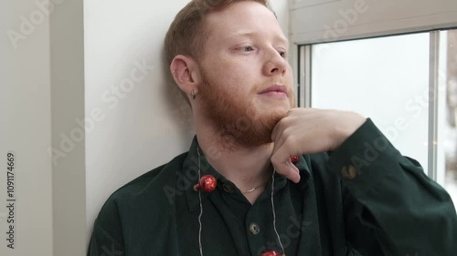 Portrait of an attractive red-haired, bearded guy sitting on the windowsill.The guy has a creative Christmas garland around his neck.New Year and Christmas concept.