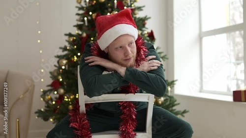 Portrait of a young red-haired man in a Santa hat sitting on a chair in front of a Christmas tree. New Year and Christmas concept.