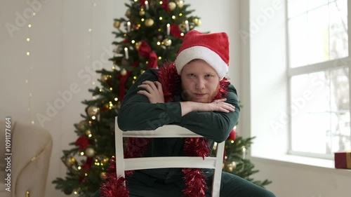 Portrait of a young red-haired man in a Santa hat sitting on a chair in front of a Christmas tree. New Year and Christmas concept.