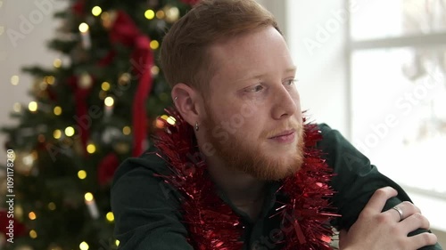 Attractive red-haired, bearded man is sitting on a chair in front of a Christmas tree in a bright living room. Portrait of a serious brutal young guy.New Year and Christmas concept.