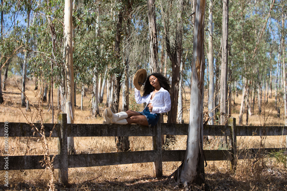 Fototapeta premium woman, young, beautiful, Latin, brunette with curly hair dressed in white shirt, denim skirt, white boots and hat, sitting with her legs stretched out on the fence of the eucalyptus forest.