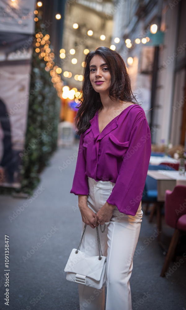 Young beautiful brunette woman in a pink jacket and white pants posing on the street. Portrait of a beautiful young woman in a purple blouse and white pants posing in an outdoor cafe on the street