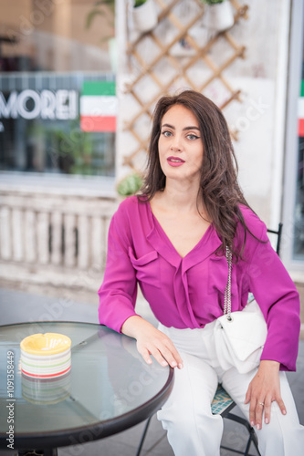 Beautiful young brunette woman in a purple blouse and white pants posing outdoor leans against a pillar .Portrait of a beautiful young brunette woman in a purple jacket posing 
