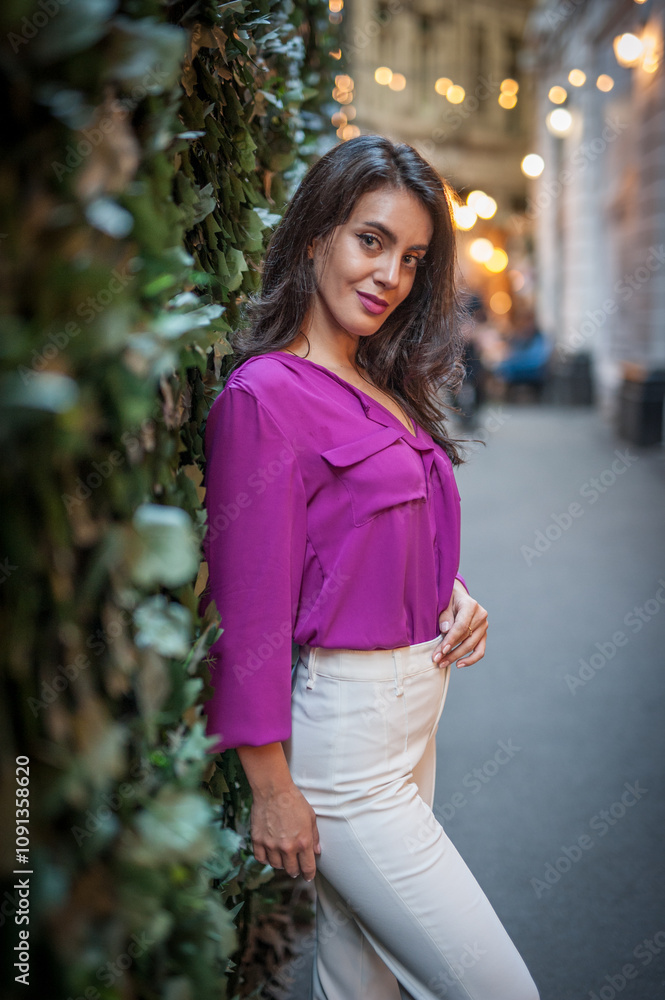 Young beautiful brunette woman in a pink jacket and white pants posing on the street. Portrait of a beautiful young woman in a purple blouse and white pants posing in an outdoor cafe on the street
