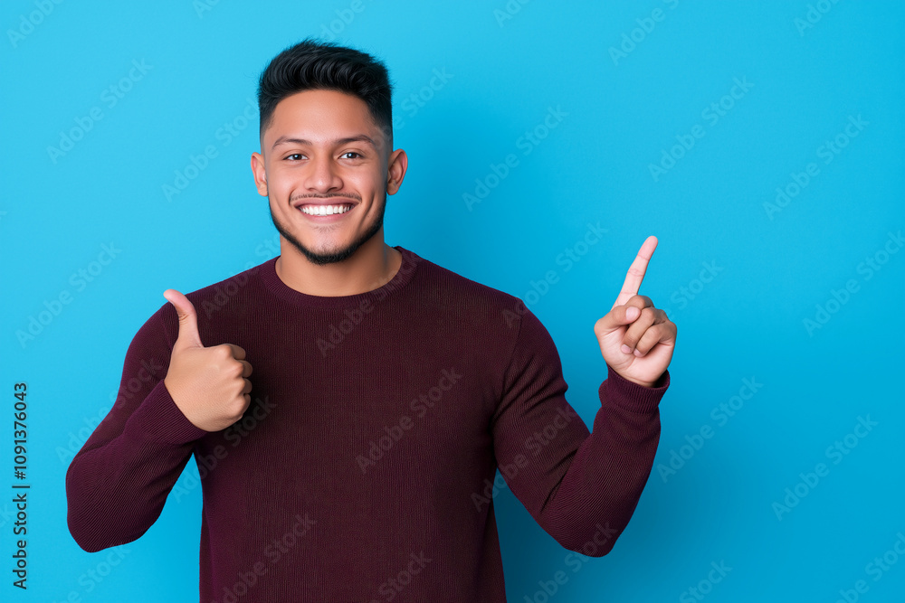 Fototapeta premium young man with cheerful expression is smiling and pointing upwards against blue background, wearing maroon sweater