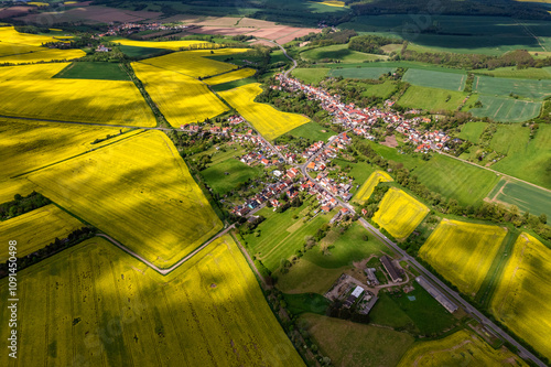 Obraz na płótnie Top view of a field of yellow rapeseed flowers with a small town in the background. The sky is cloudy and the sun is shining through the clouds. Germany. w salonie