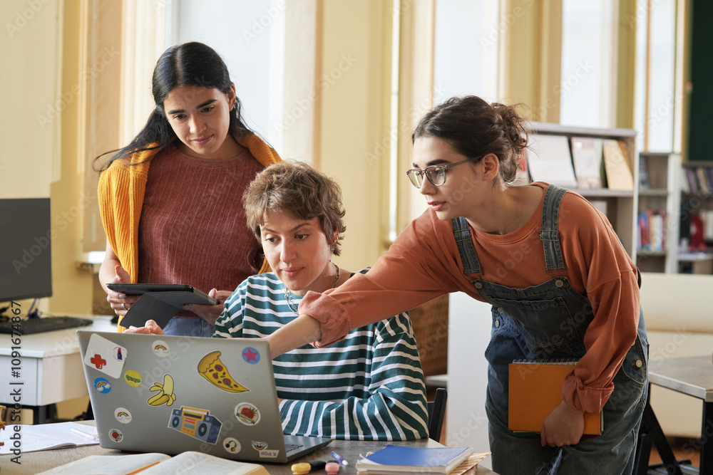 Obraz premium Three students are collaborating on a group project while using a laptop and tablet in classroom environment with bookshelves and windows