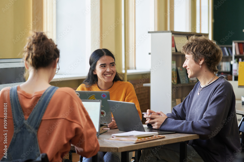 Fototapeta premium Group of students sitting at table in university library engaged in group discussion, using laptops and notebooks, with bookshelves in background