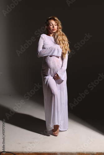 Beautiful pregnant woman in special white wet dress. Advertisement of comfortable pregnancy Happy blond pregnant girl stand in studio with studio light. Long curly hair. Black background