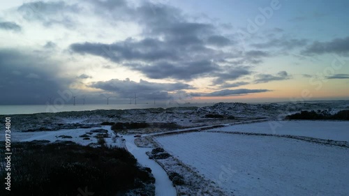Drone reveals wind turbines generating renewable energy out to sea beyond snow on sandy dunes beach on the North East coast of Scotaland