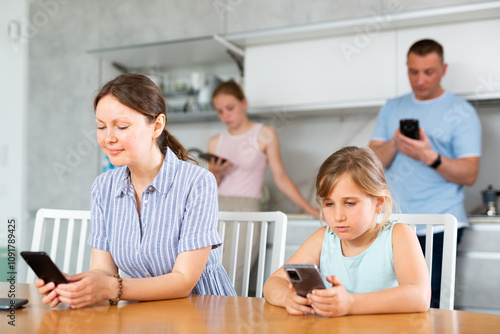 Smiling woman mom sitting near kitchen table next to younger daughter with phone in hands, playing online game, watching video recipes. Father of family and eldest daughter surf Internet in background photo